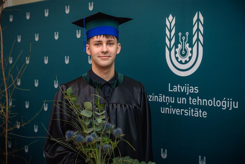 LBTU absolvents Arnolds Polis absolvēšanas tērpā ar diplomu un ziediem pie LBTU sienas ar universitātes logotipu.