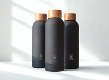 Trīs melnas ūdens pudeles ar LBTU logo un koka korģi. Gaišs fons ar loga ēnojumu. 