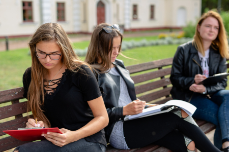 Trīs studentes sēž uz soliņa universitātes teritorijā un raksta piezīmes kladēs un planšetēs, koncentrējoties uz studiju uzdevumu svaigā gaisā.