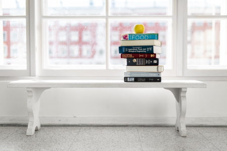 Balts sols pie liela loga, uz kura novietota grāmatu kaudze ar dzeltenu ābolu virsū; fonā gaiša, izplūdusi universitātes vide.