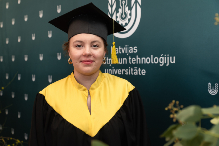 LBTU absolvente Vilma Alberinga absolventes tērpā un cepurē pozē pie zaļa banera ar LBTU logo fonā.