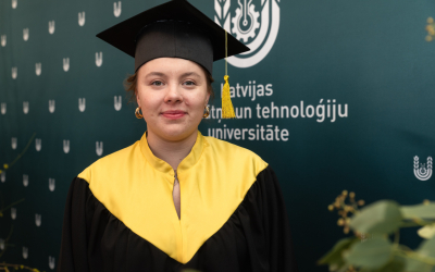LBTU absolvente Vilma Alberinga absolventes tērpā un cepurē pozē pie zaļa banera ar LBTU logo fonā.